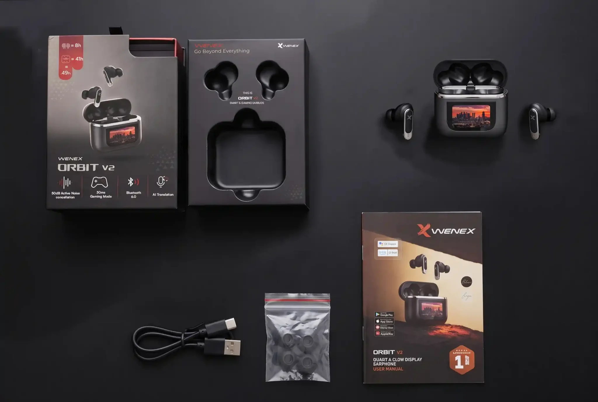 Everything in the Orbit V2 box: the earbuds in their open charging case, the retail box and inner tray, a USB-C cable, spare ear tips and the printed guide, laid out on a dark studio surface.